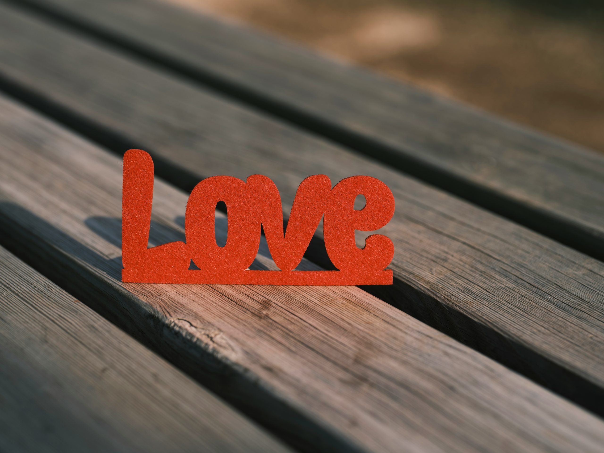 A red cutout of the word Love stands on a wooden surface, sunlight casting shadows, evoking the warmth and hope found in The Certainty of Chance, with a softly blurred background.