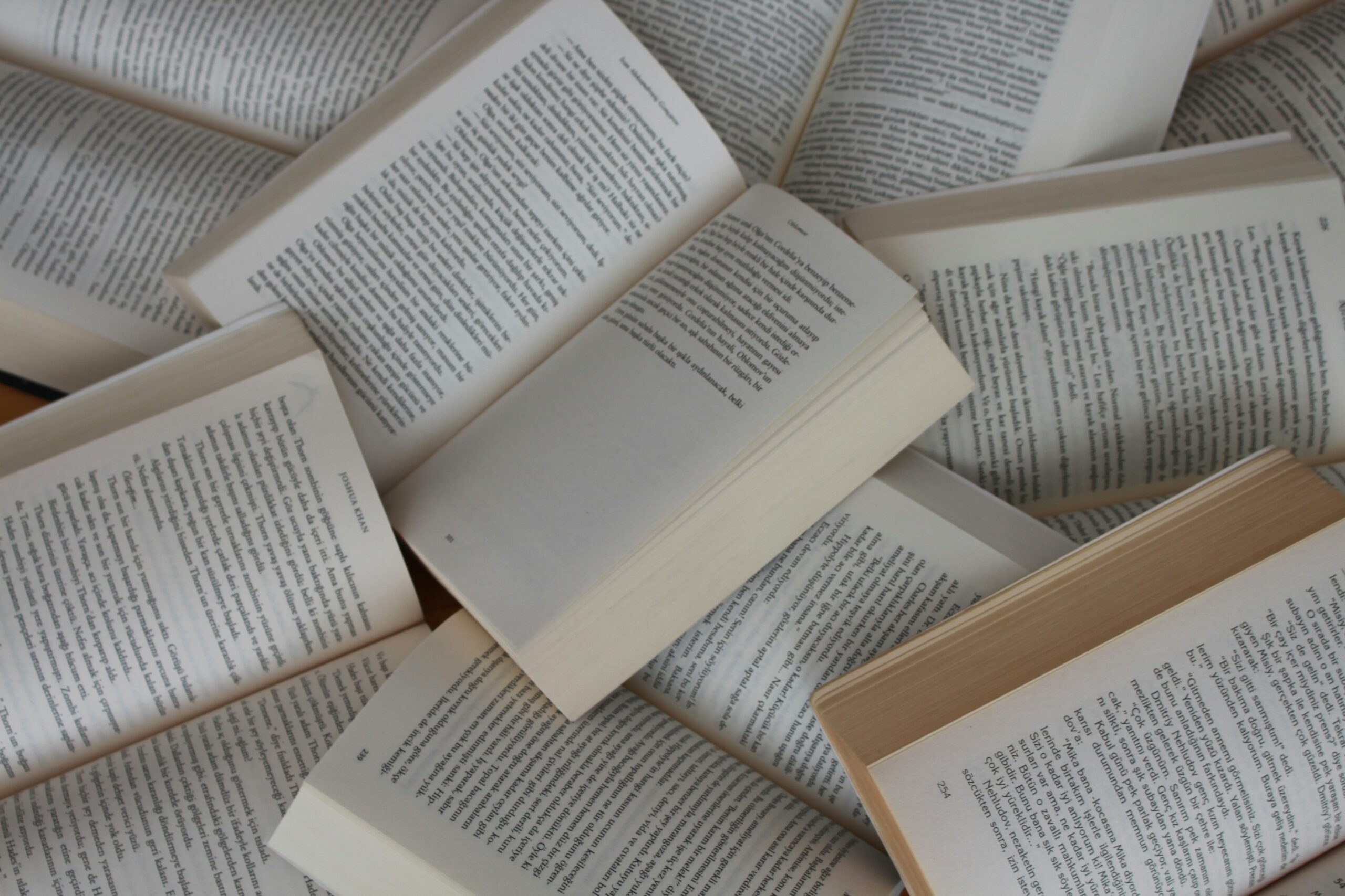 Several open books are scattered and overlapping, showing pages filled with dense text in a serif font. The books are arranged haphazardly, creating a layered, textured pattern of printed words.
