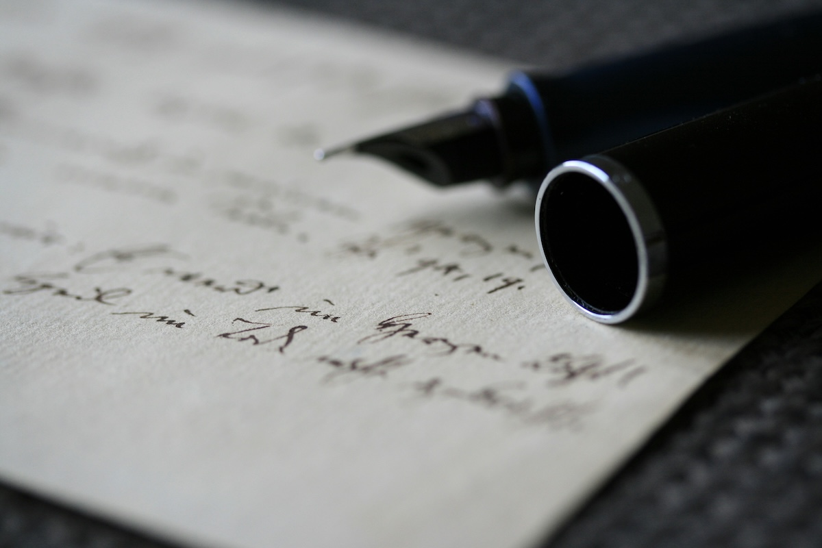 A close-up of a black fountain pen resting on a handwritten letter, featuring elegant cursive on cream-colored paper. The focus is on the pen’s nib and the script, hinting at secrets behind The Trouble with Heroes.